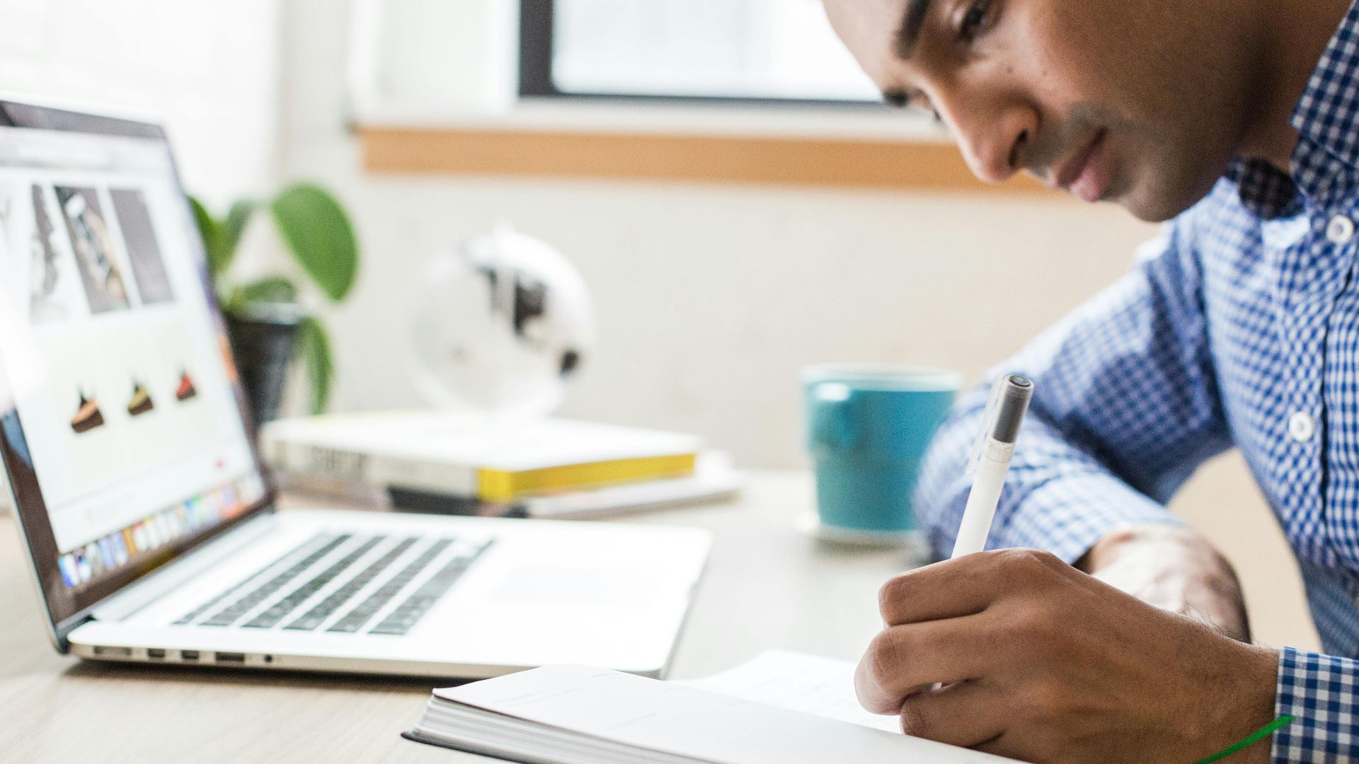 Man focused on writing in a notebook at an office desk with a laptop and coffee cup.