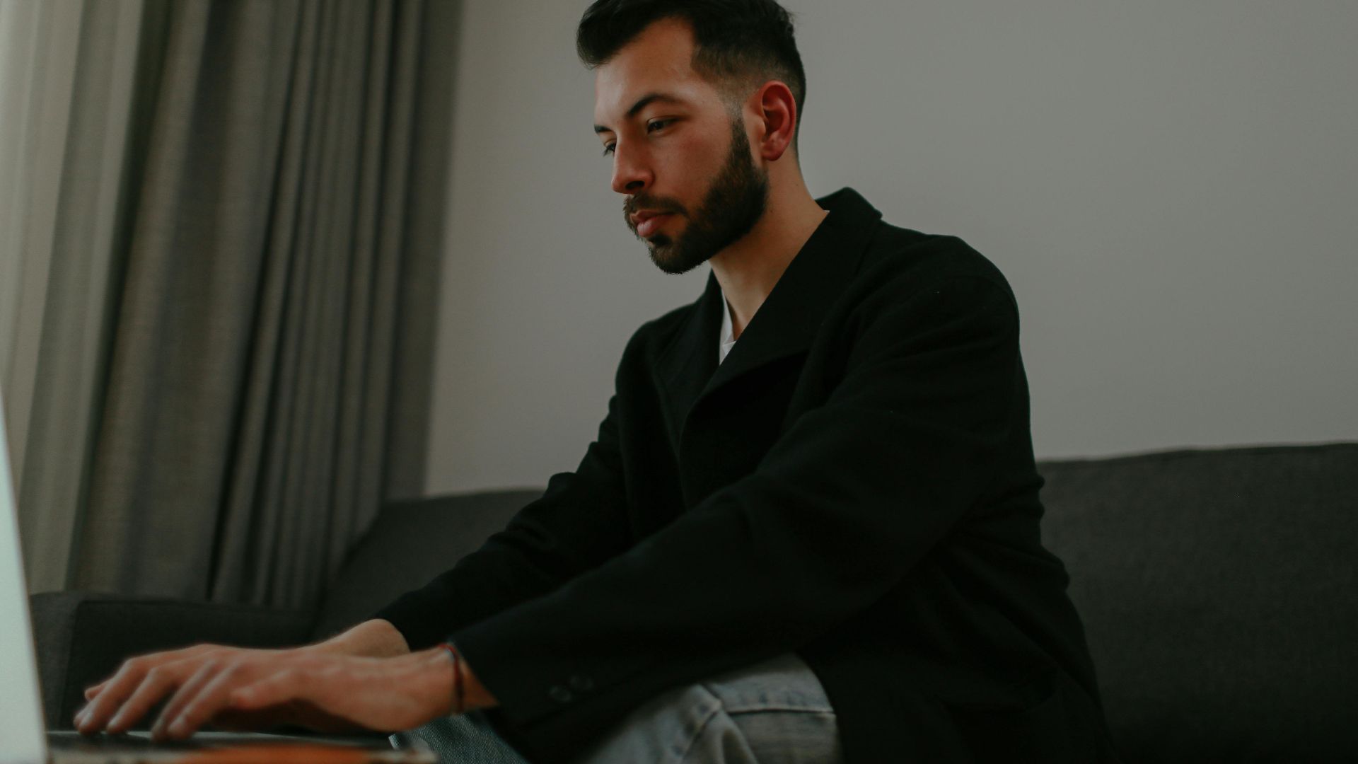 Man in a black blazer sitting on a sofa, focused on typing on his laptop indoors.