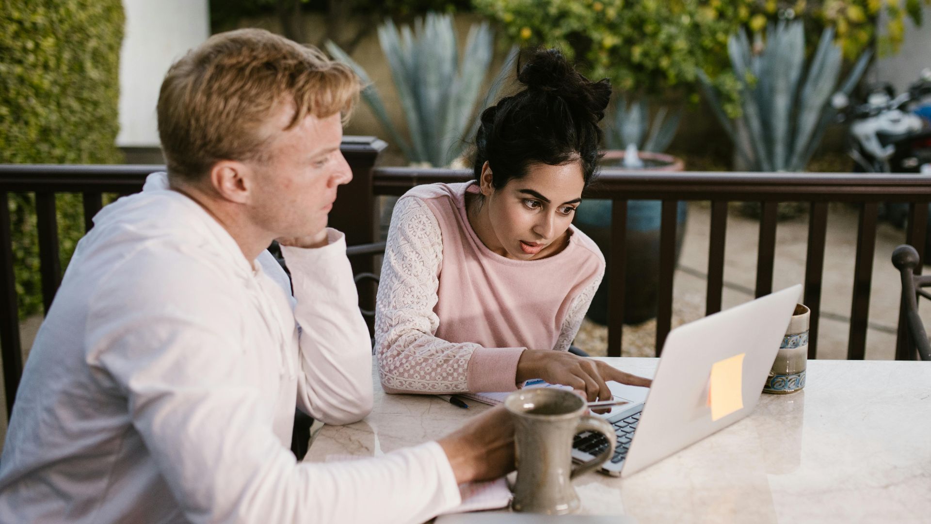 Two adults collaborating on a laptop, seated outdoors with coffee on the table.