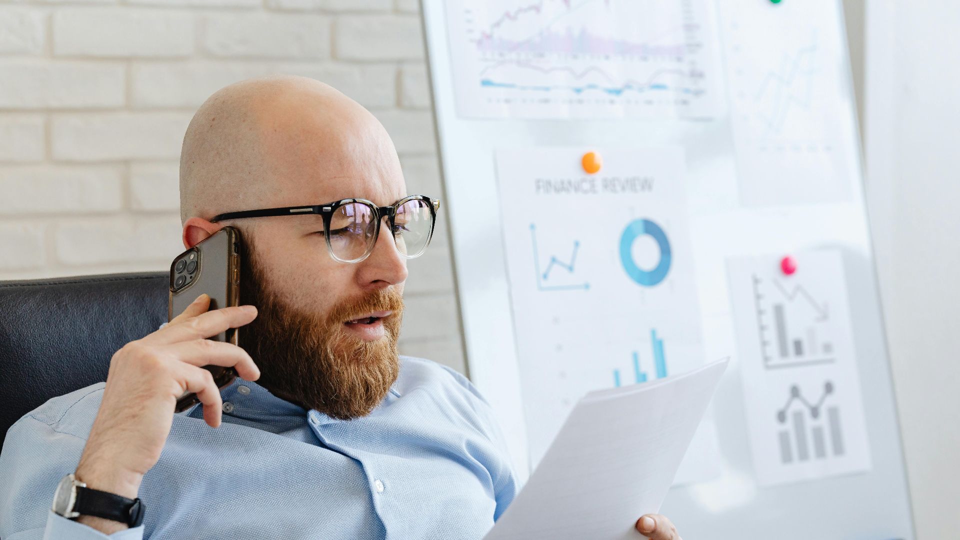 Bald man with beard holding smartphone and papers, reviewing financial charts in office.