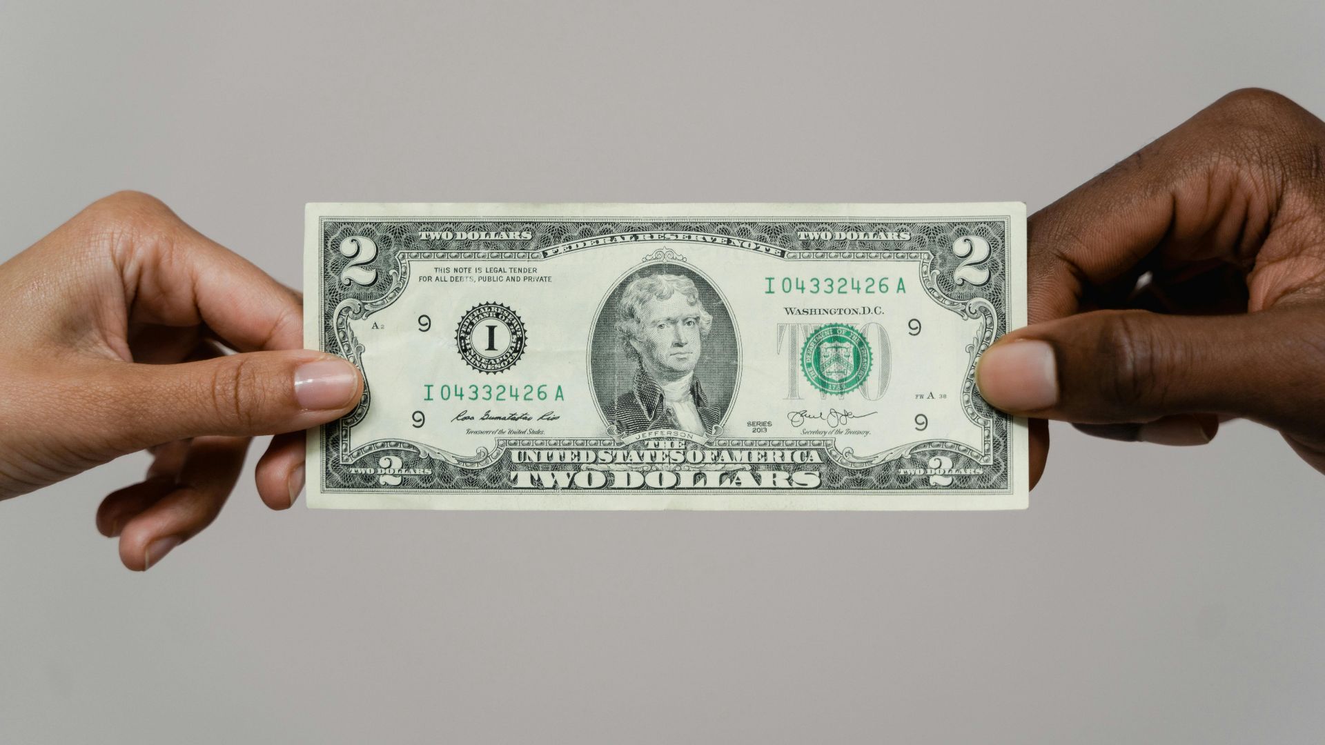 Close-up of two hands exchanging a two-dollar bill on a neutral background.