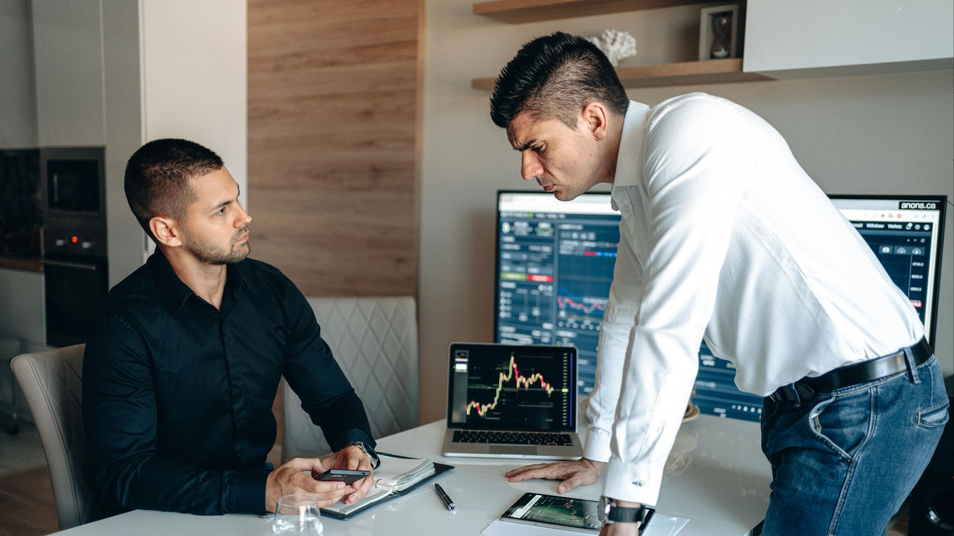Two businessmen analyzing stock market data on laptops and tablets in an office environment.