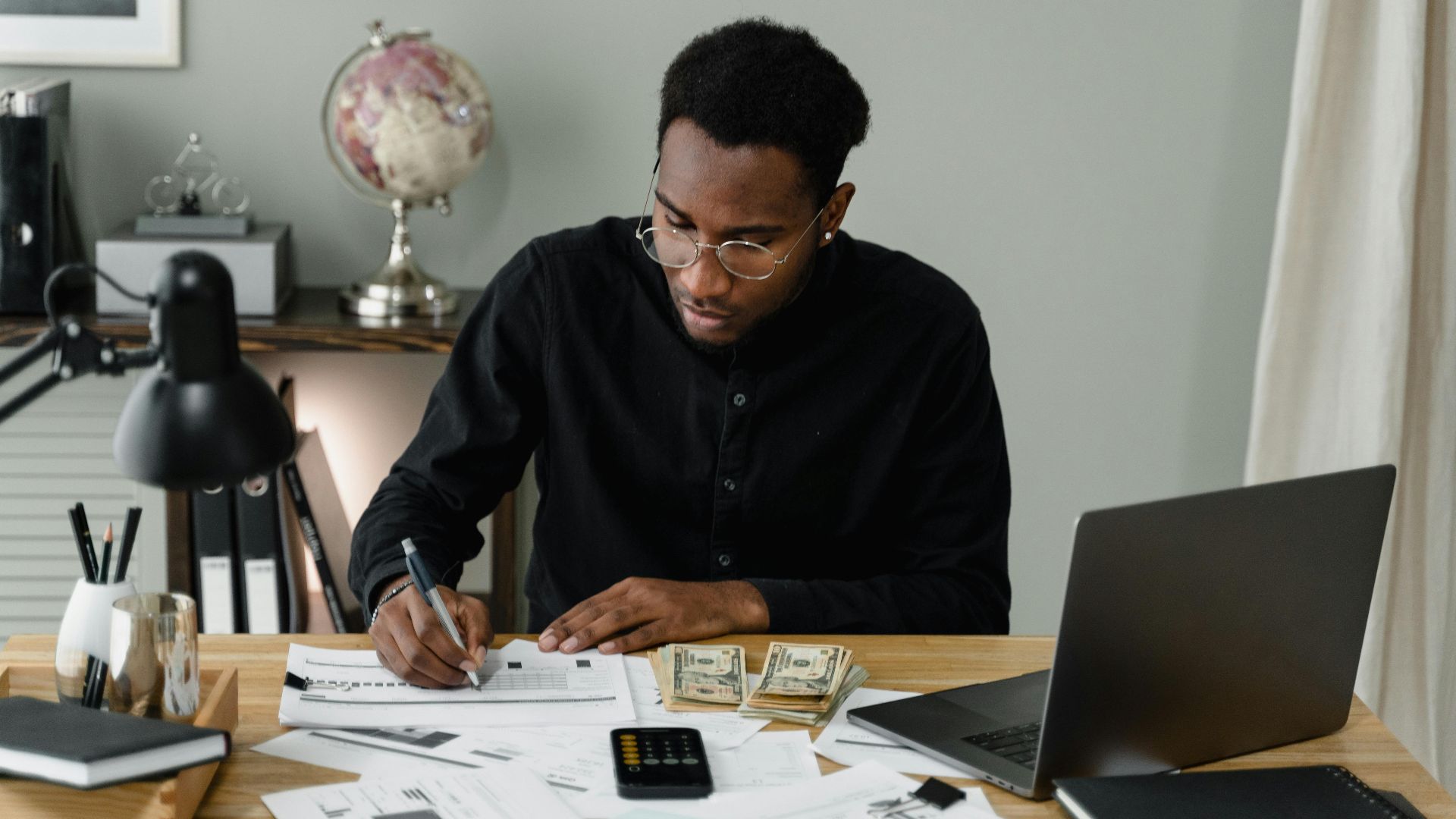 An adult man calculates expenses, using a laptop and documents at a desk in a home office setting.