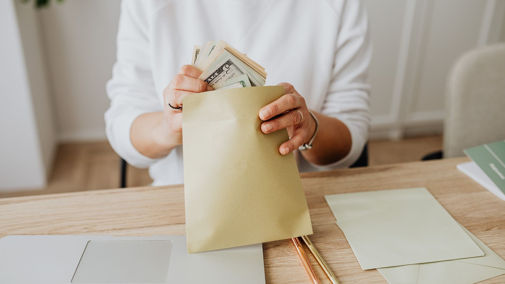Close-up of person holding envelopes with cash at a wooden desk indoors.