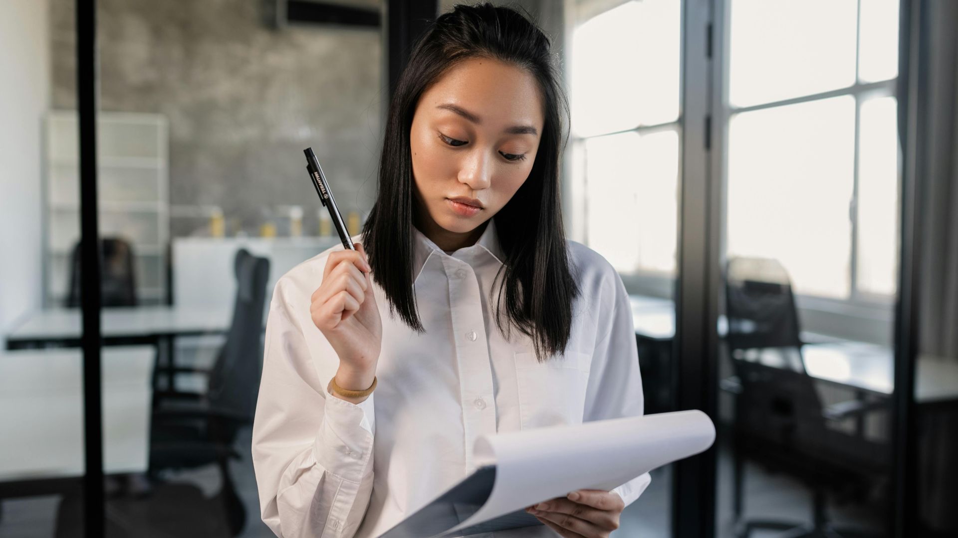 Confident Asian woman in office reviewing documents with pen.