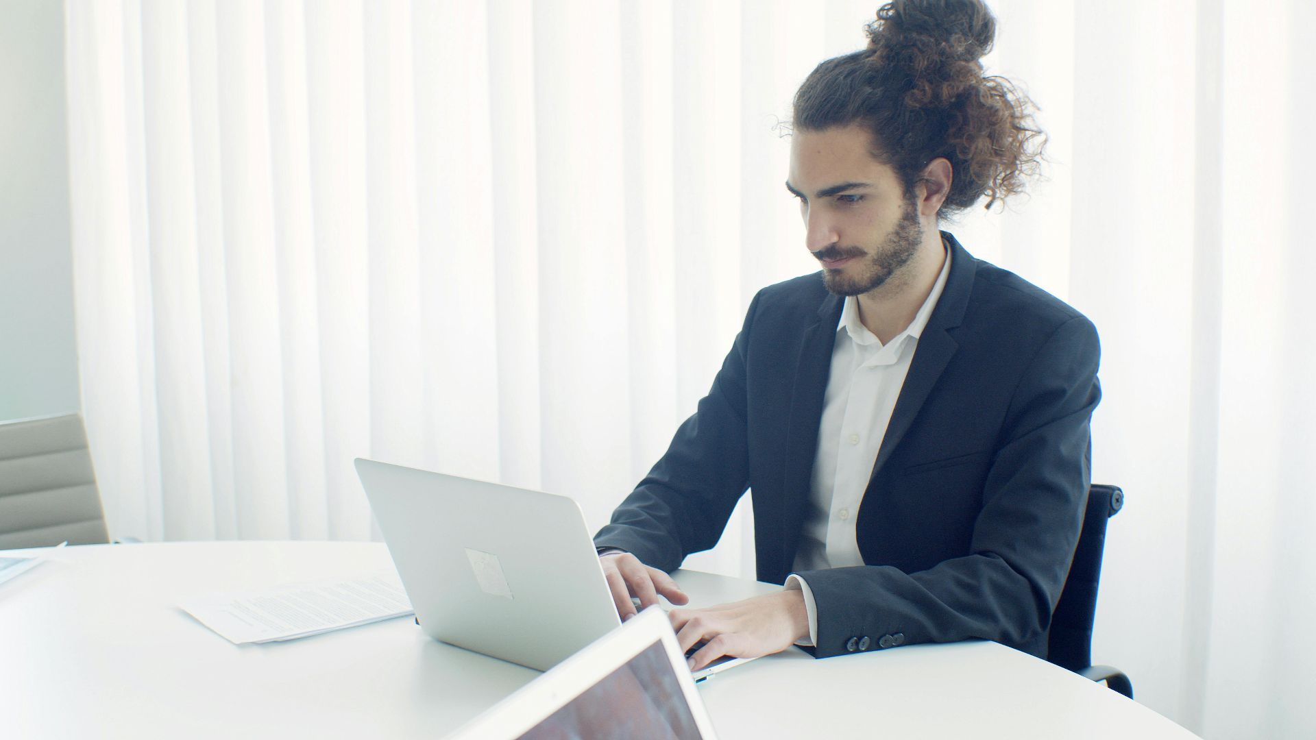 Businessman with hair bun typing on laptop in bright modern office setting.