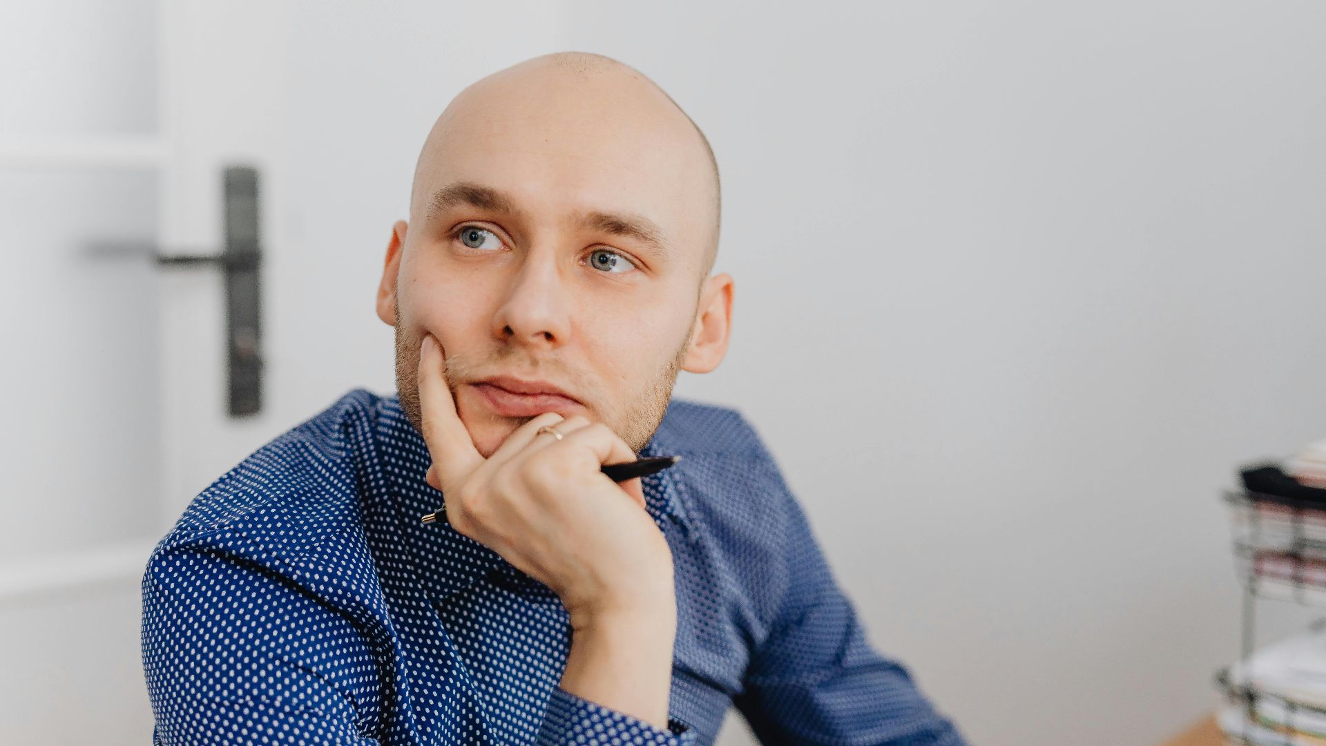 Bald man in blue shirt thoughtfully holding a pen indoors, gazing into distance.