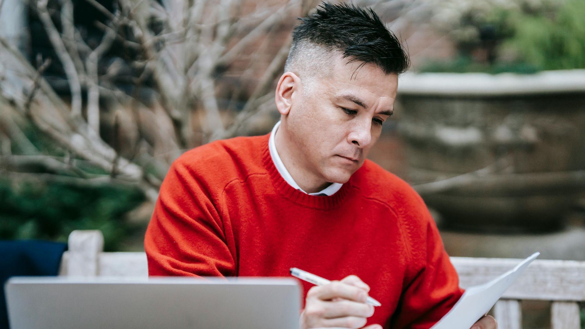 A man in a red sweater concentrating on paperwork while working remotely with a laptop outdoors.