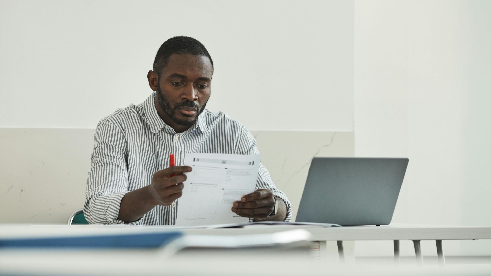 An adult man attentively grading test papers at his workspace with a laptop.