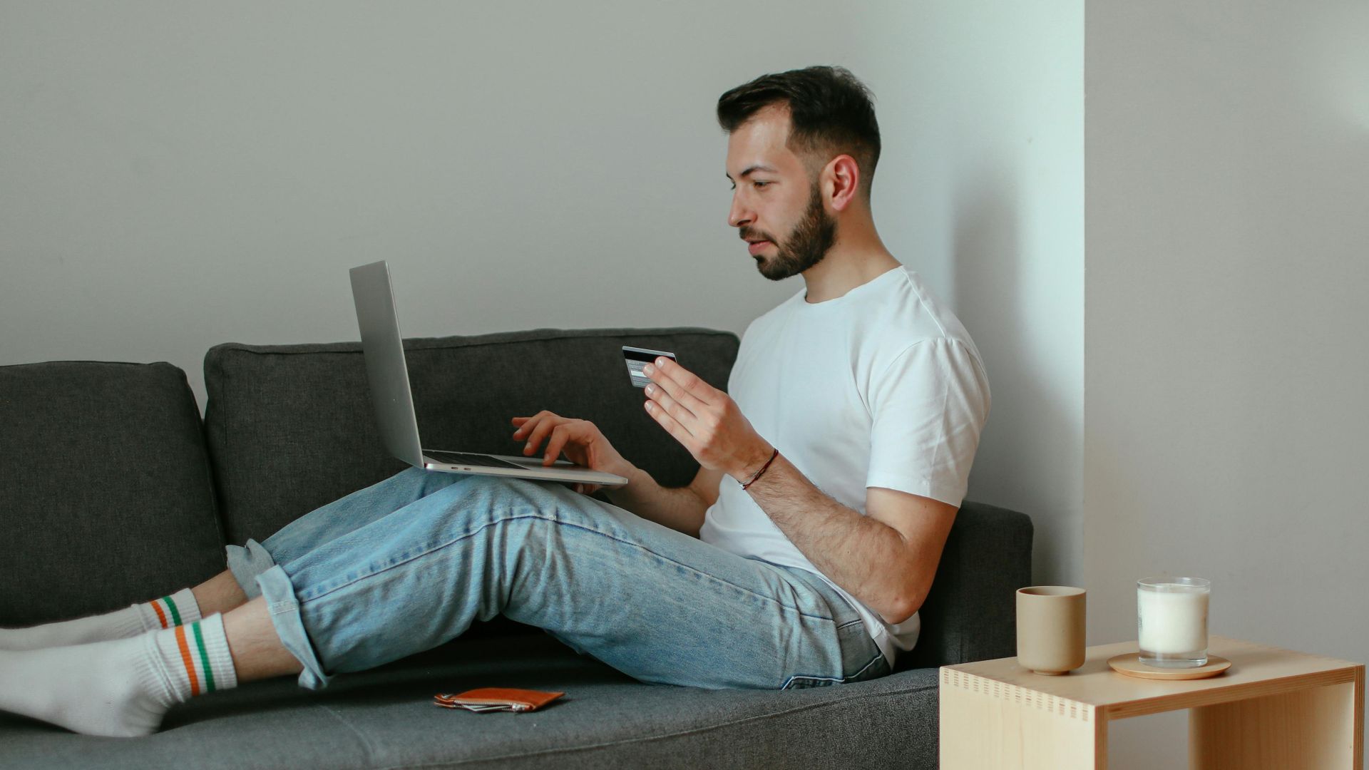 Adult man using laptop for online shopping on sofa, holding credit card.