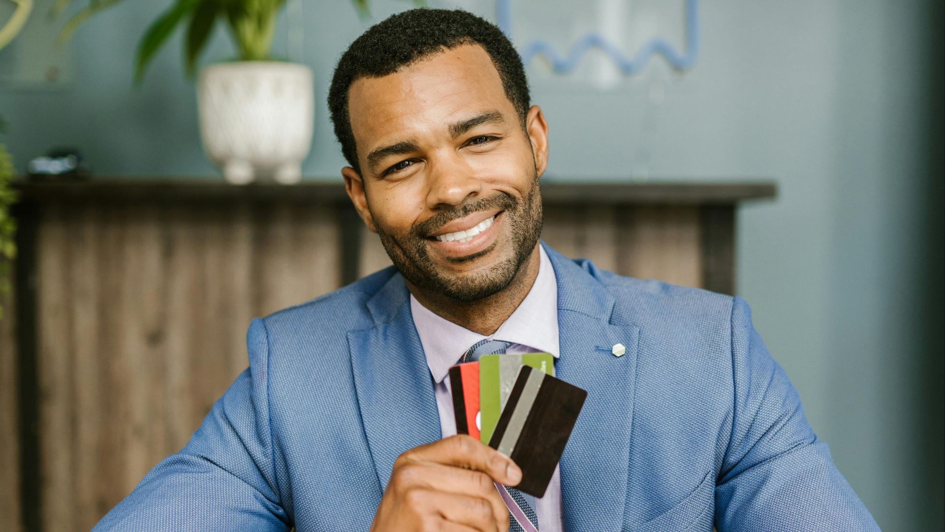 A confident businessman in a blue suit holds and displays credit cards while smiling at a desk.