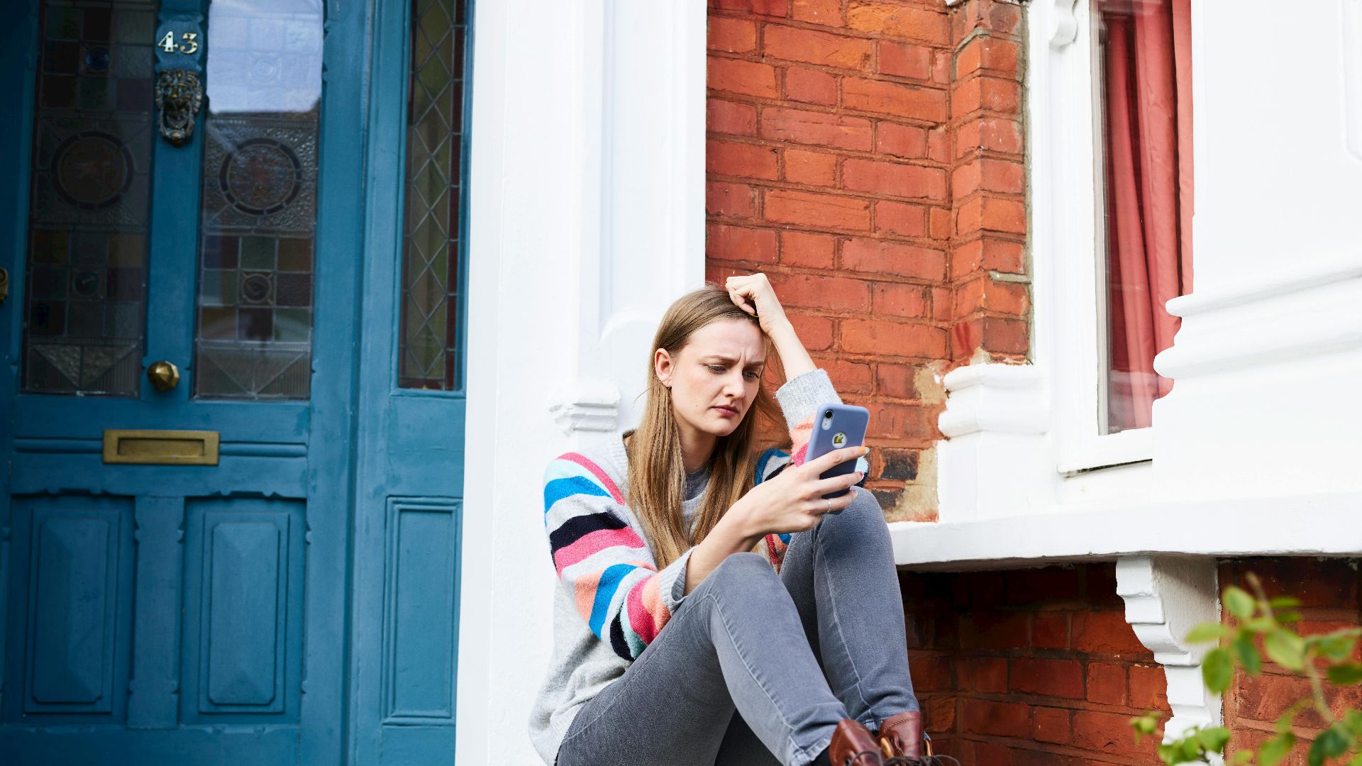 Young woman using phone outside red brick house with blue door, looking thoughtful.