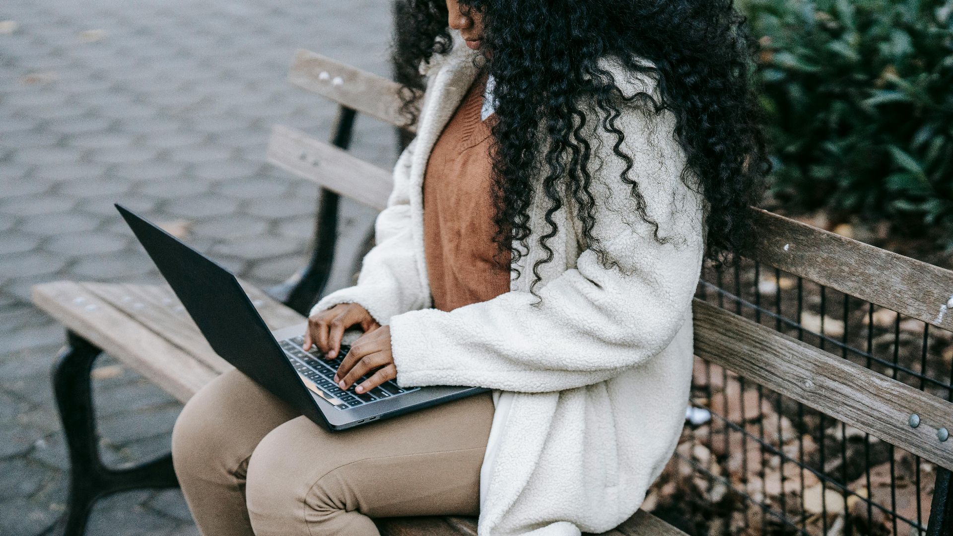 Crop concentrated African American female in warm jacket working on modern netbook and sitting on wooden bench in spring city park