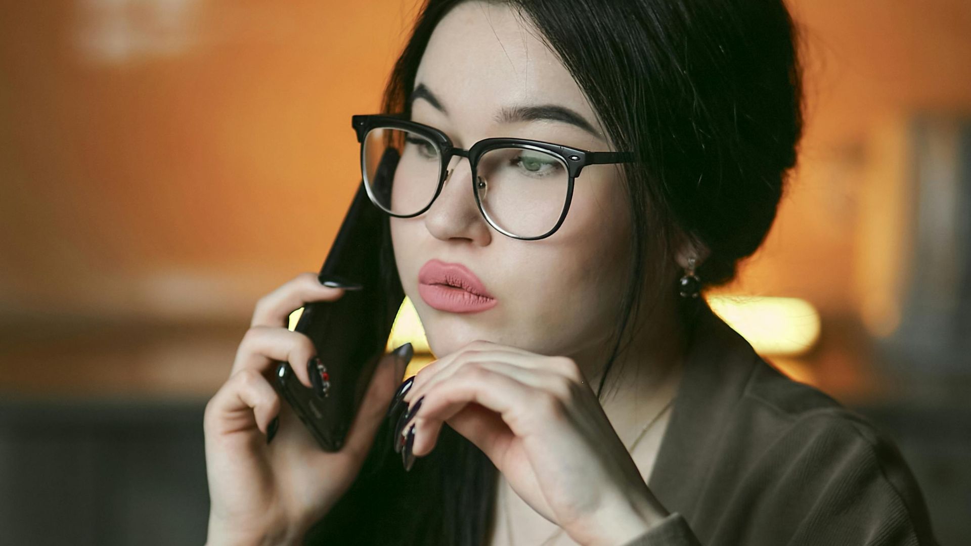 A woman with eyeglasses engaged in a phone call indoors, focused and thoughtful.