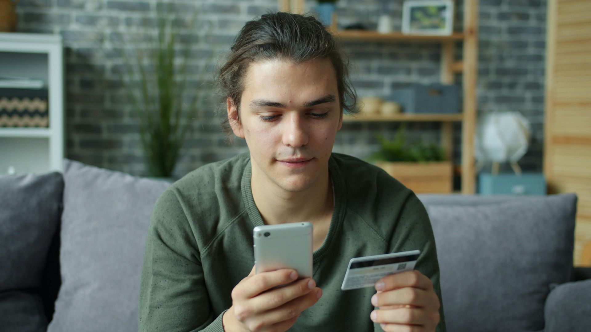 A young man using a smartphone and credit card for online shopping at home.