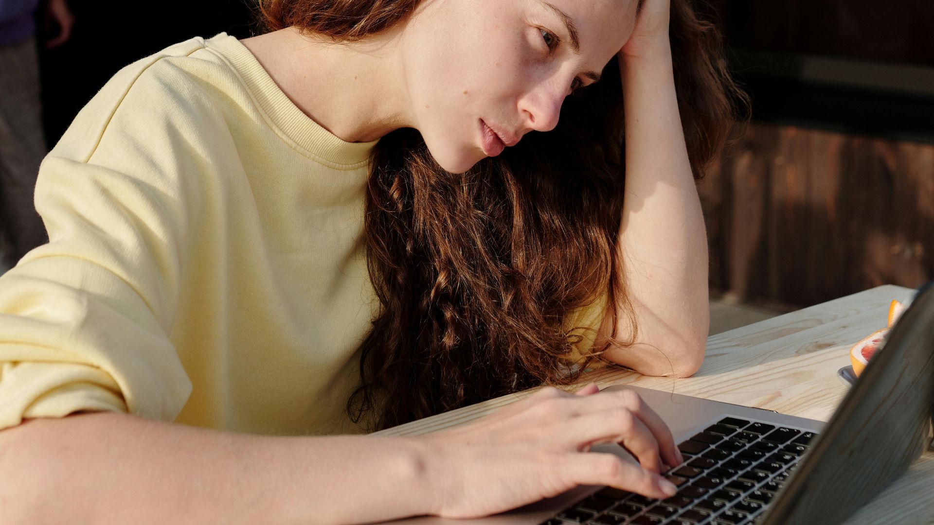 A young woman with long hair works outdoors on a laptop, appearing focused and thoughtful.