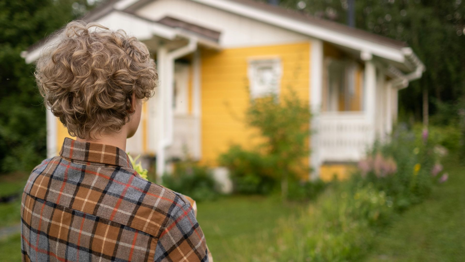 Teen with curly hair in plaid shirt stands facing a charming yellow house amidst lush greenery.
