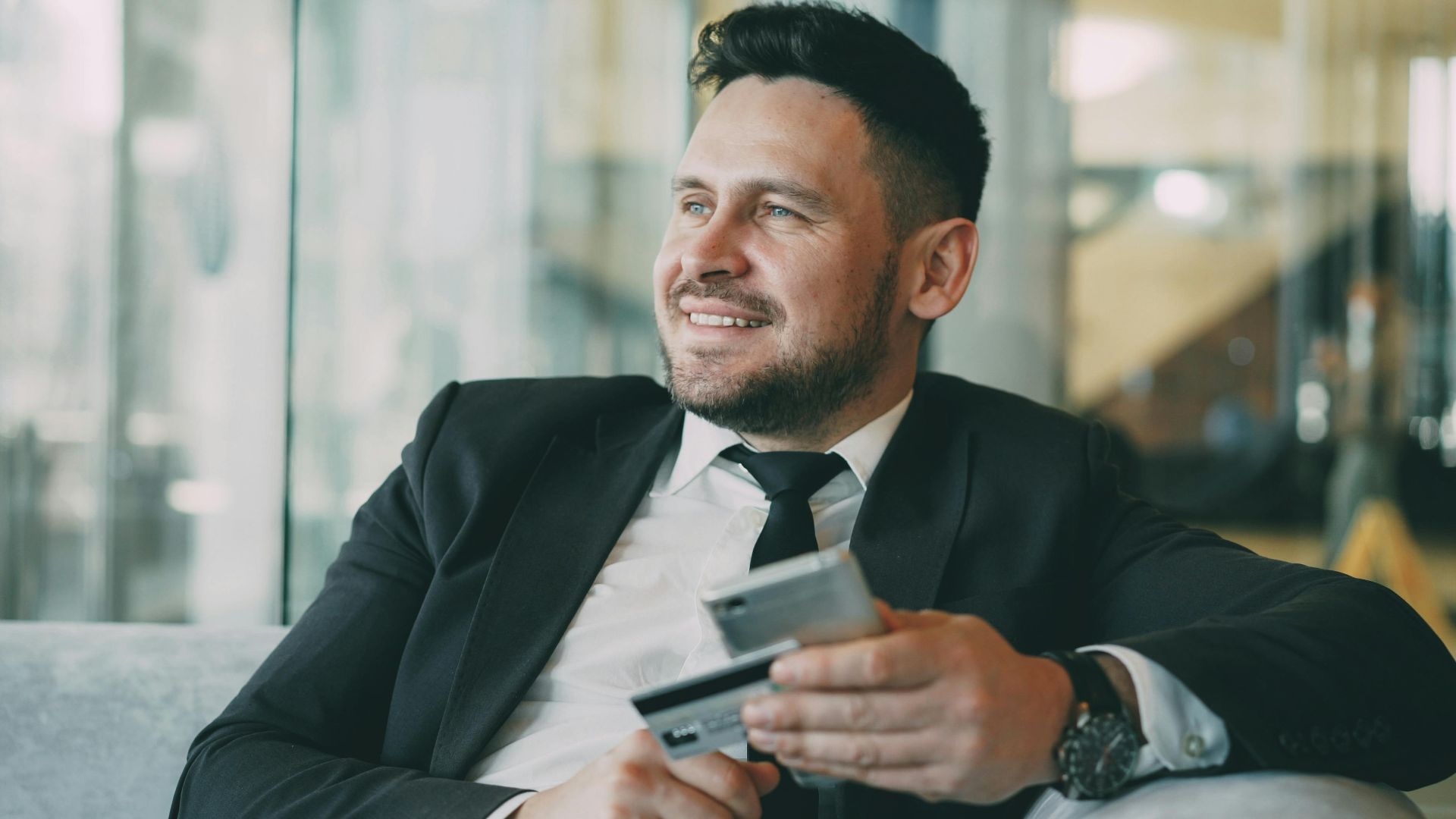 Smiling businessman in a suit holds a credit card and phone indoors, exuding confidence.