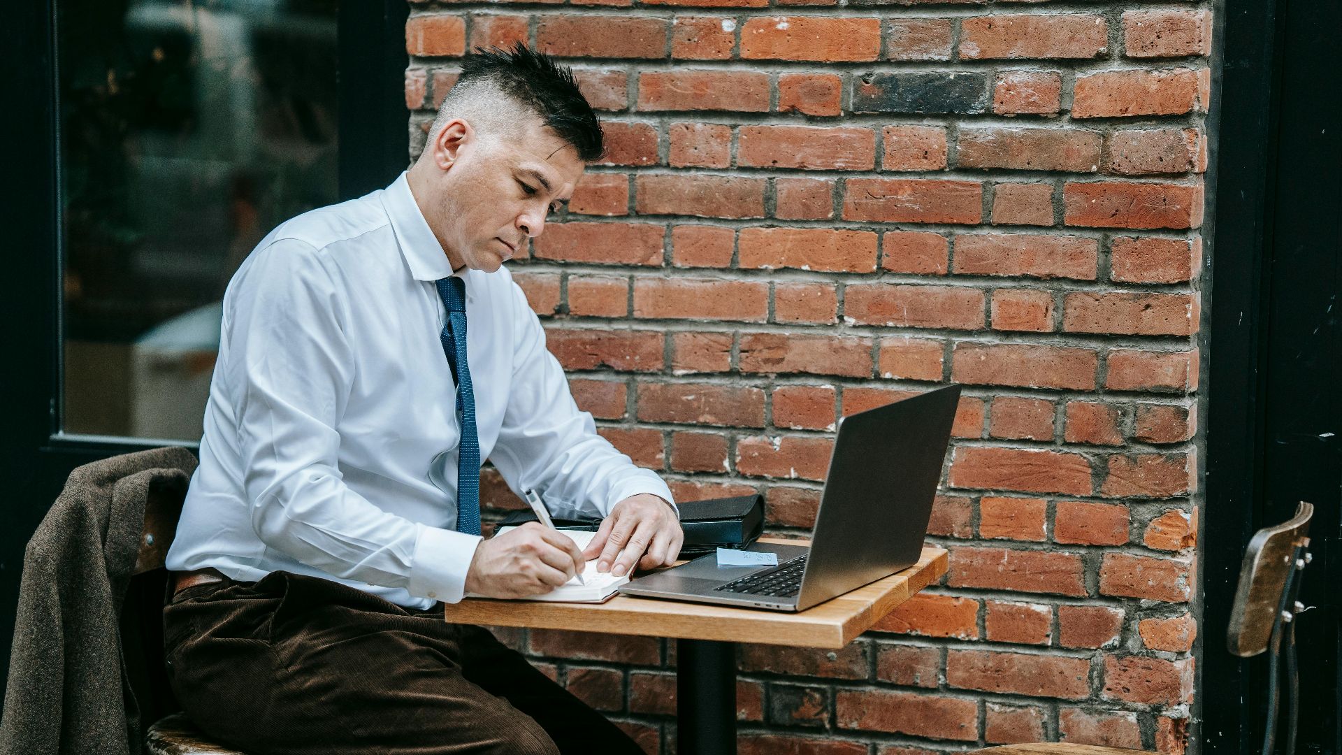 Adult man in corporate attire working on laptop at a cafe table outdoors.