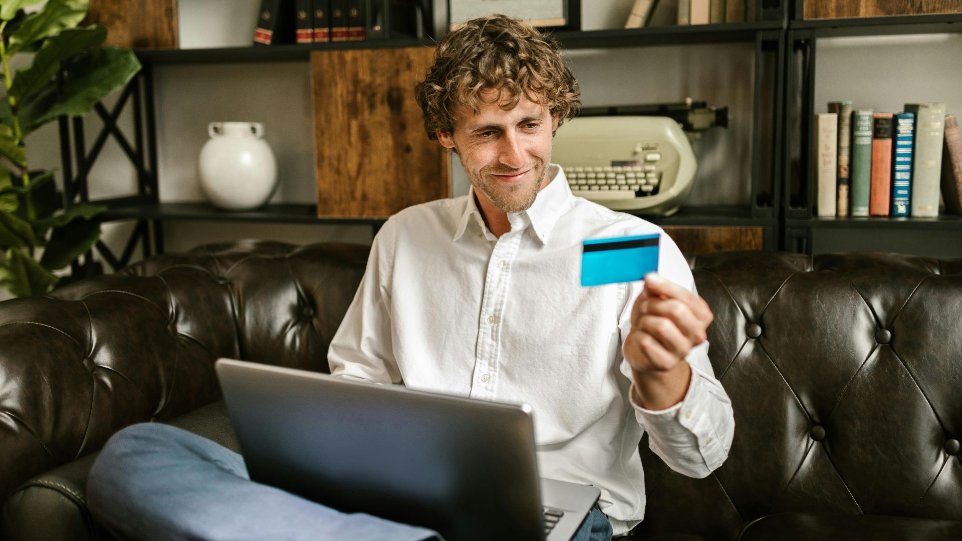 A man sits on a sofa, smiling while holding a credit card with a laptop on his lap, indoors.