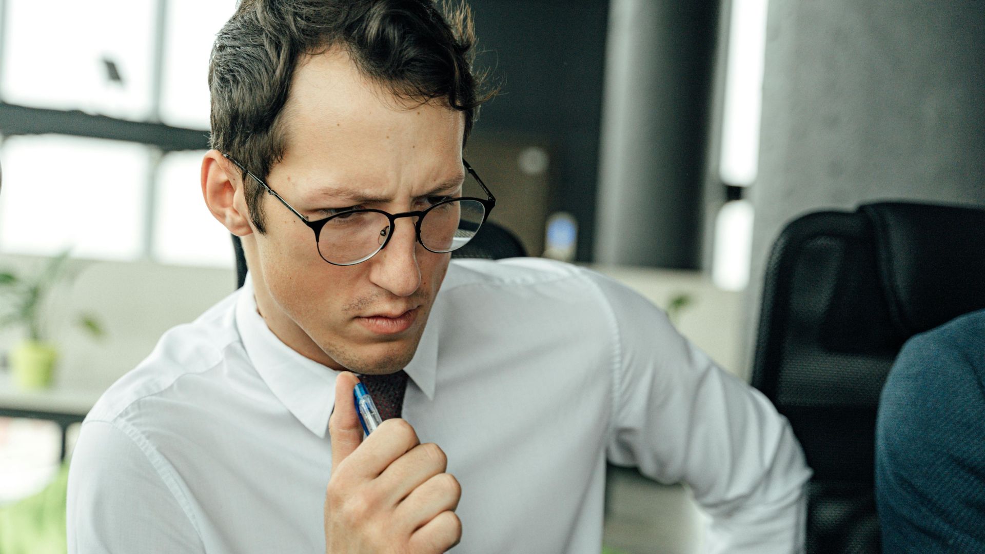 Man in white shirt thinking deeply at office desk with pen.