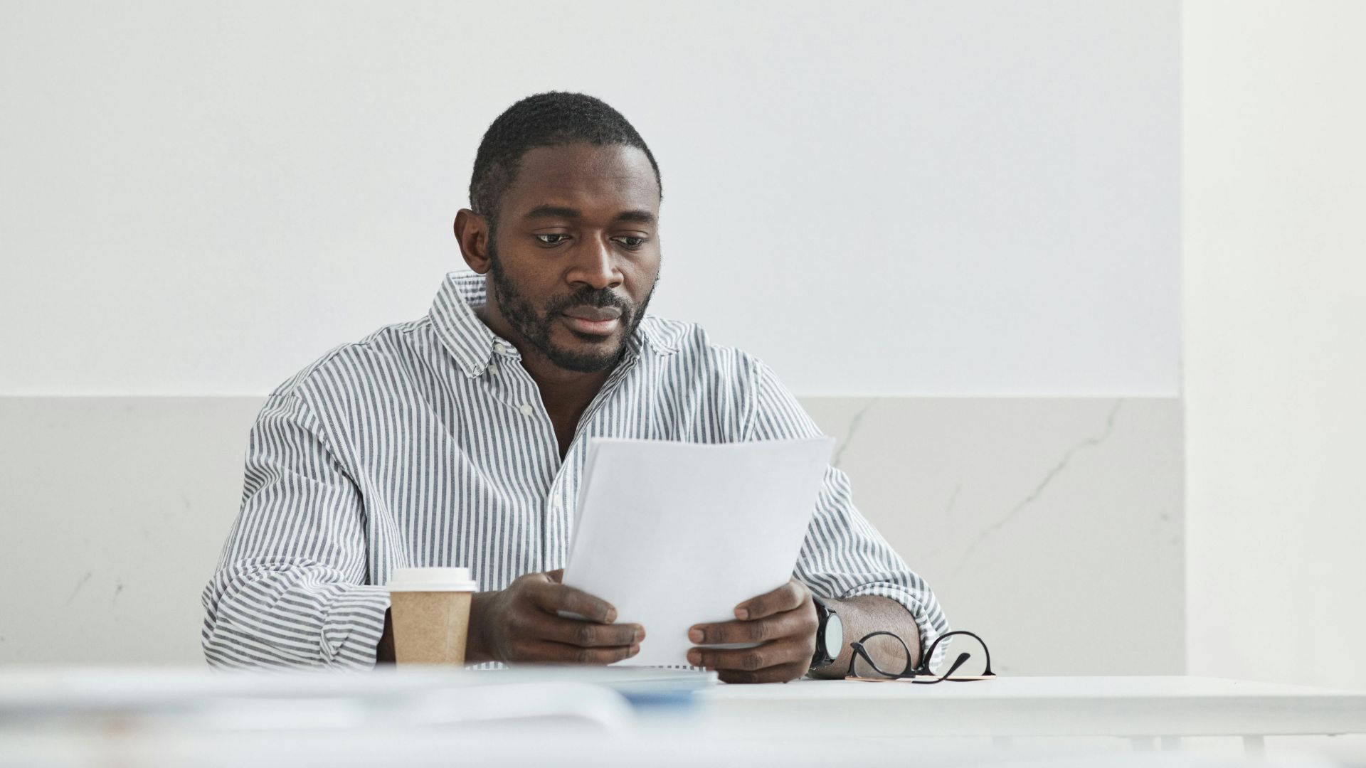 African American man sitting indoors, reading papers with a coffee cup nearby.