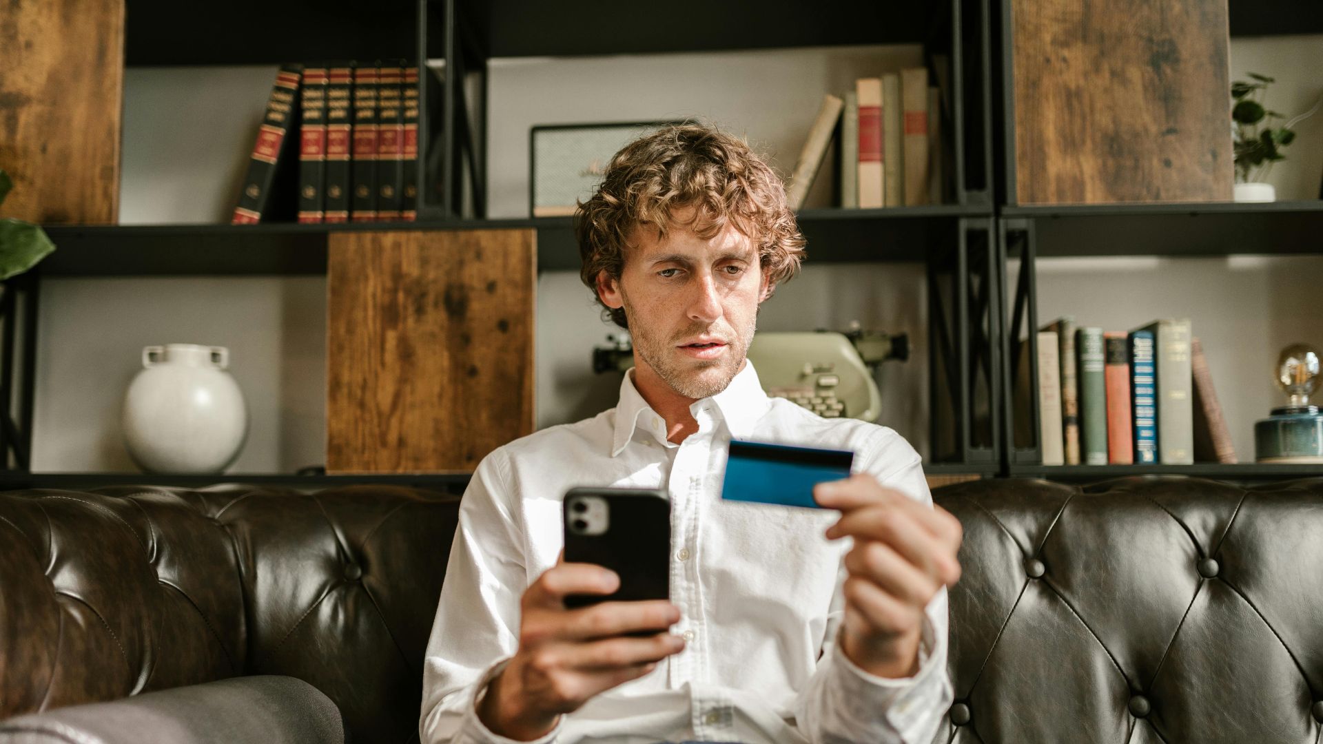 A man sitting on a leather sofa using a smartphone and credit card for online shopping.
