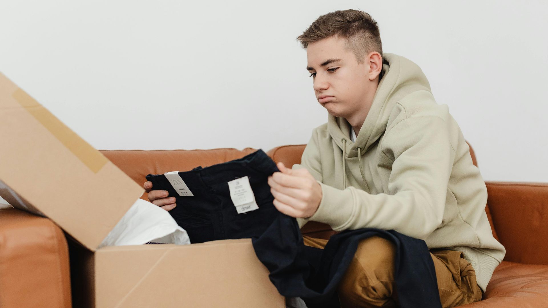 Teenager on couch opening package, unhappy with clothing. Indoors, casual setting.