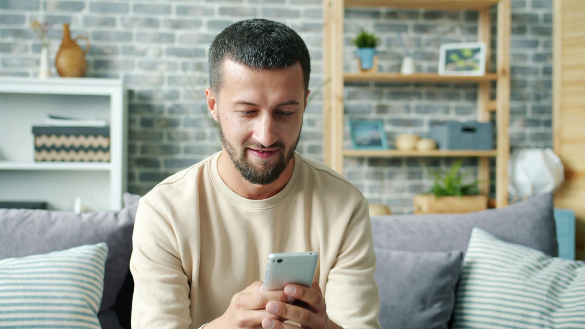 Casually dressed man using smartphone while sitting on a cozy sofa in a modern living room.