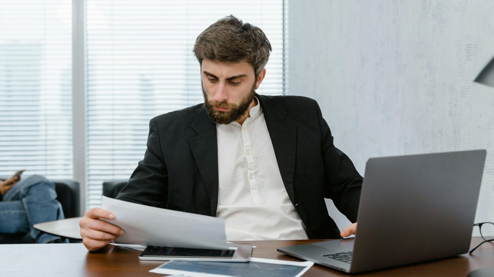 Focused businessman reviews financial reports at office desk with laptop.
