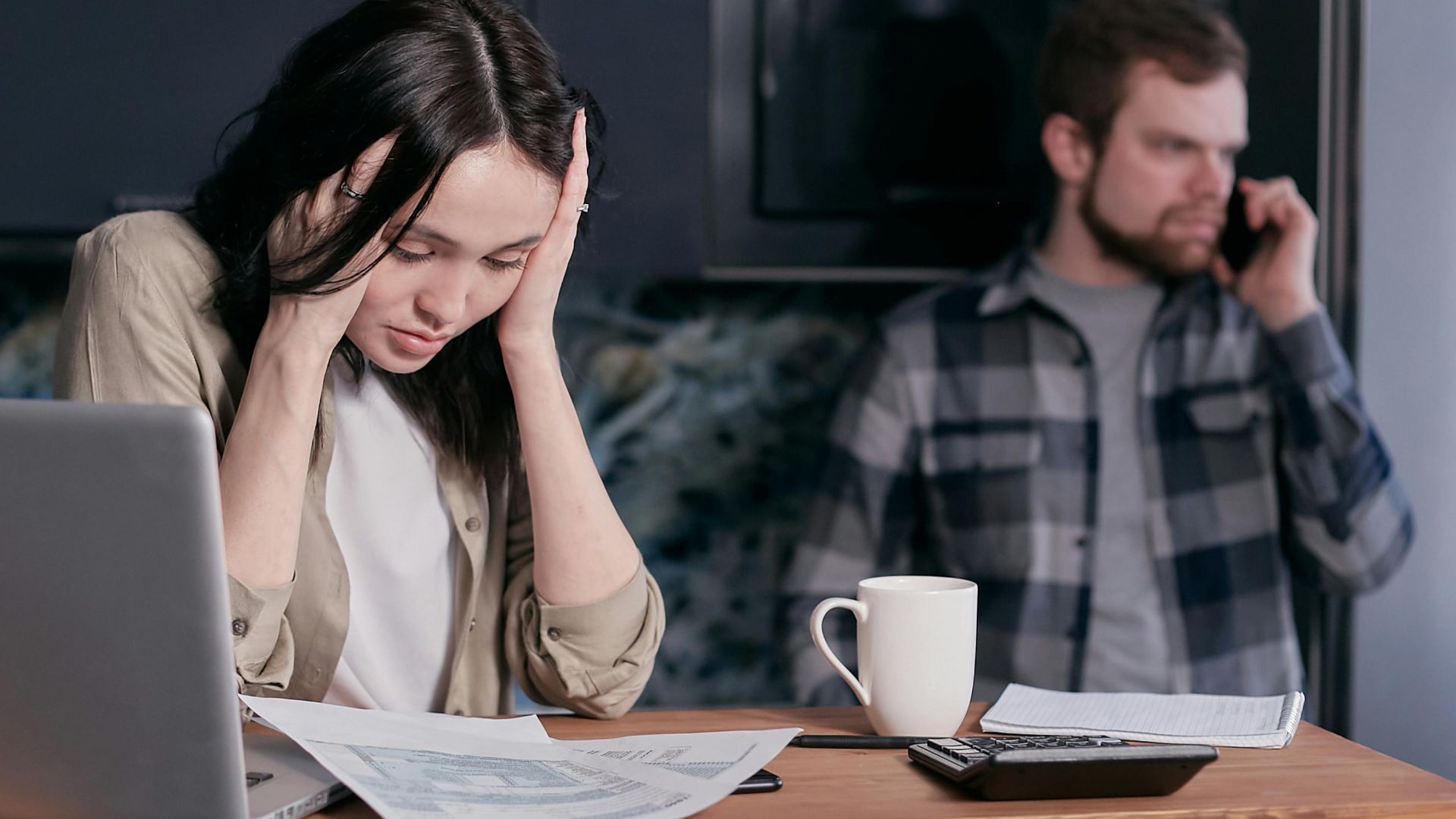 A stressed woman holds her head while working at a desk with documents and a laptop.
