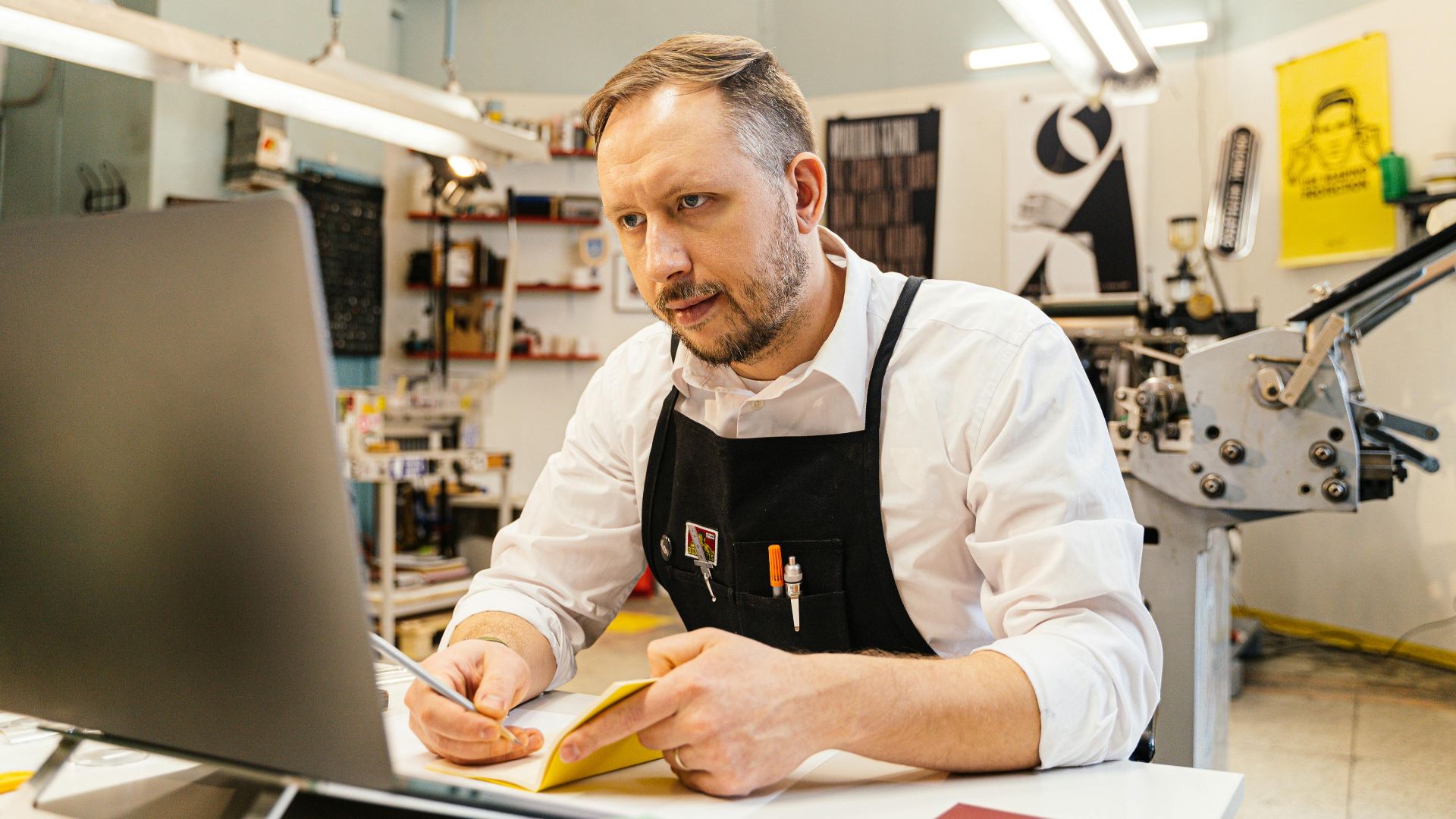 A dedicated man working at his computer within a busy print shop space, focusing intently.