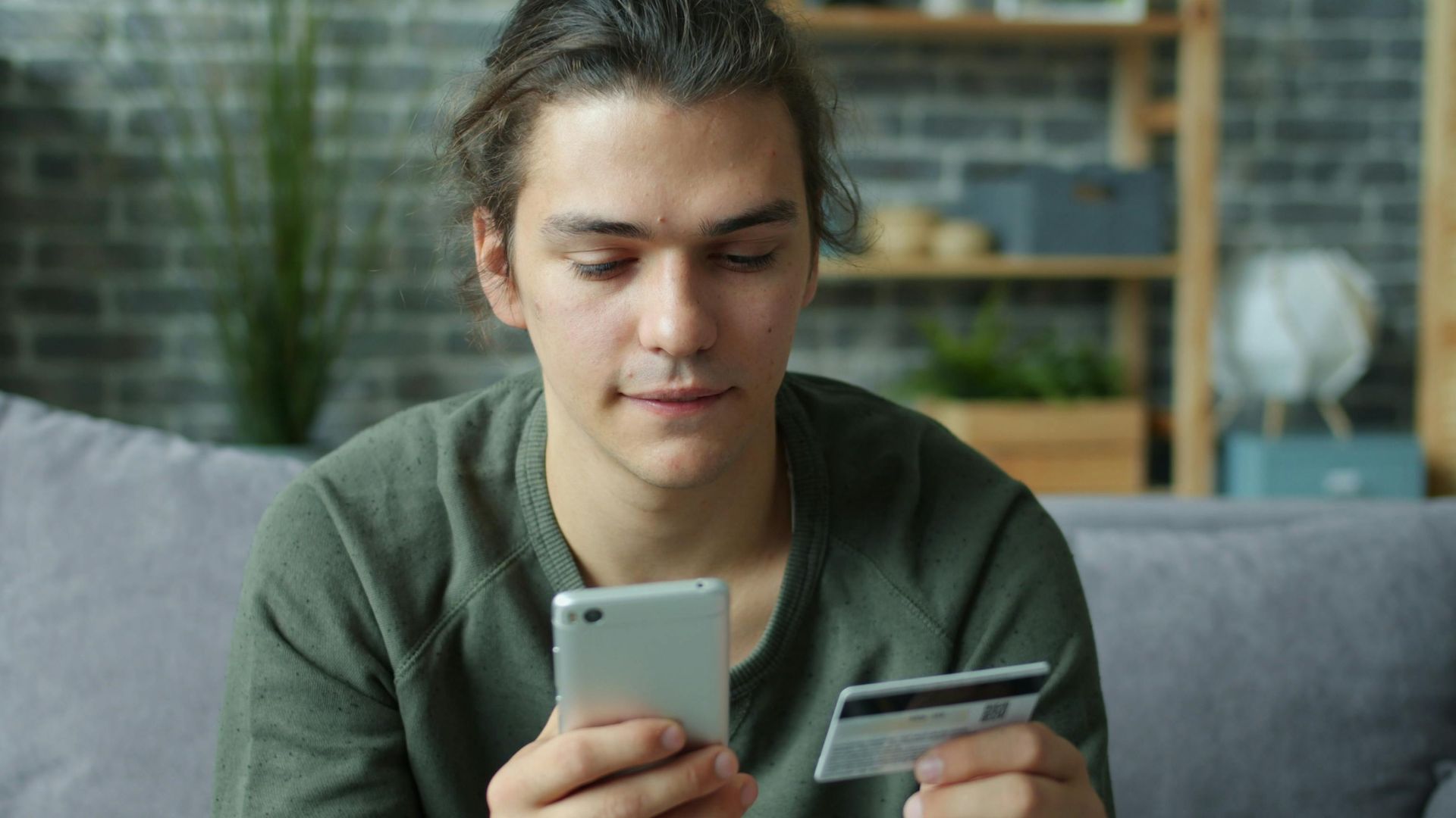 A young man using a smartphone and credit card for online shopping at home.