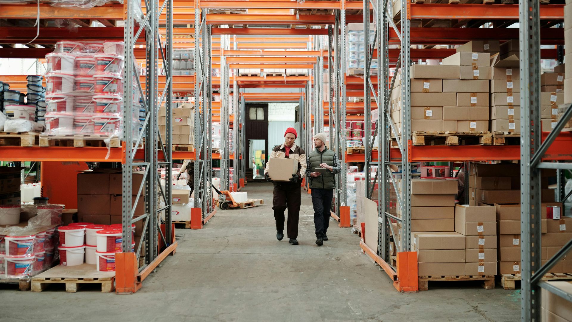 Two workers handle a package in a spacious warehouse surrounded by shelves stocked with boxes and products.