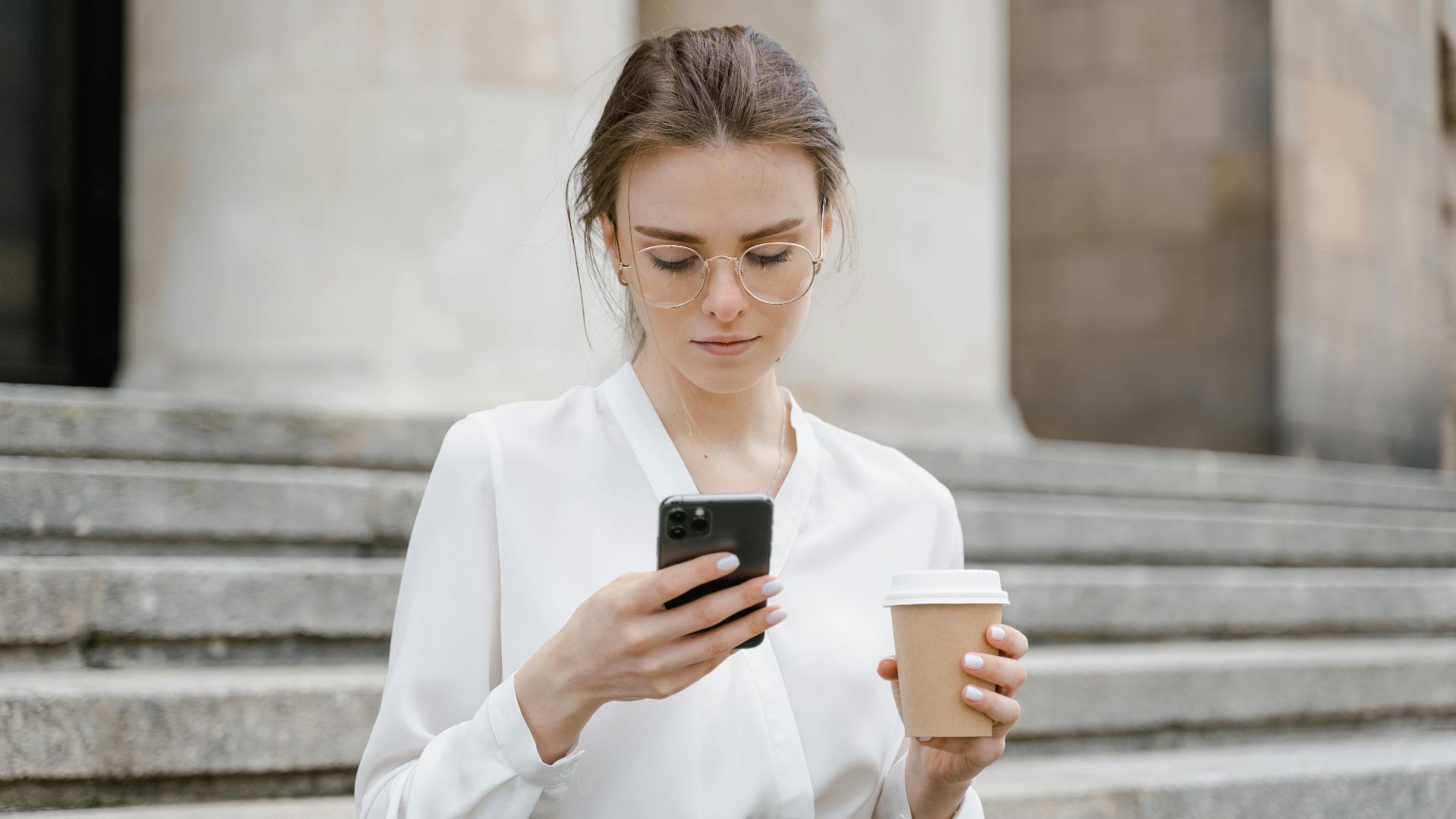 A young woman in glasses texting on her phone while holding a coffee cup outdoors.