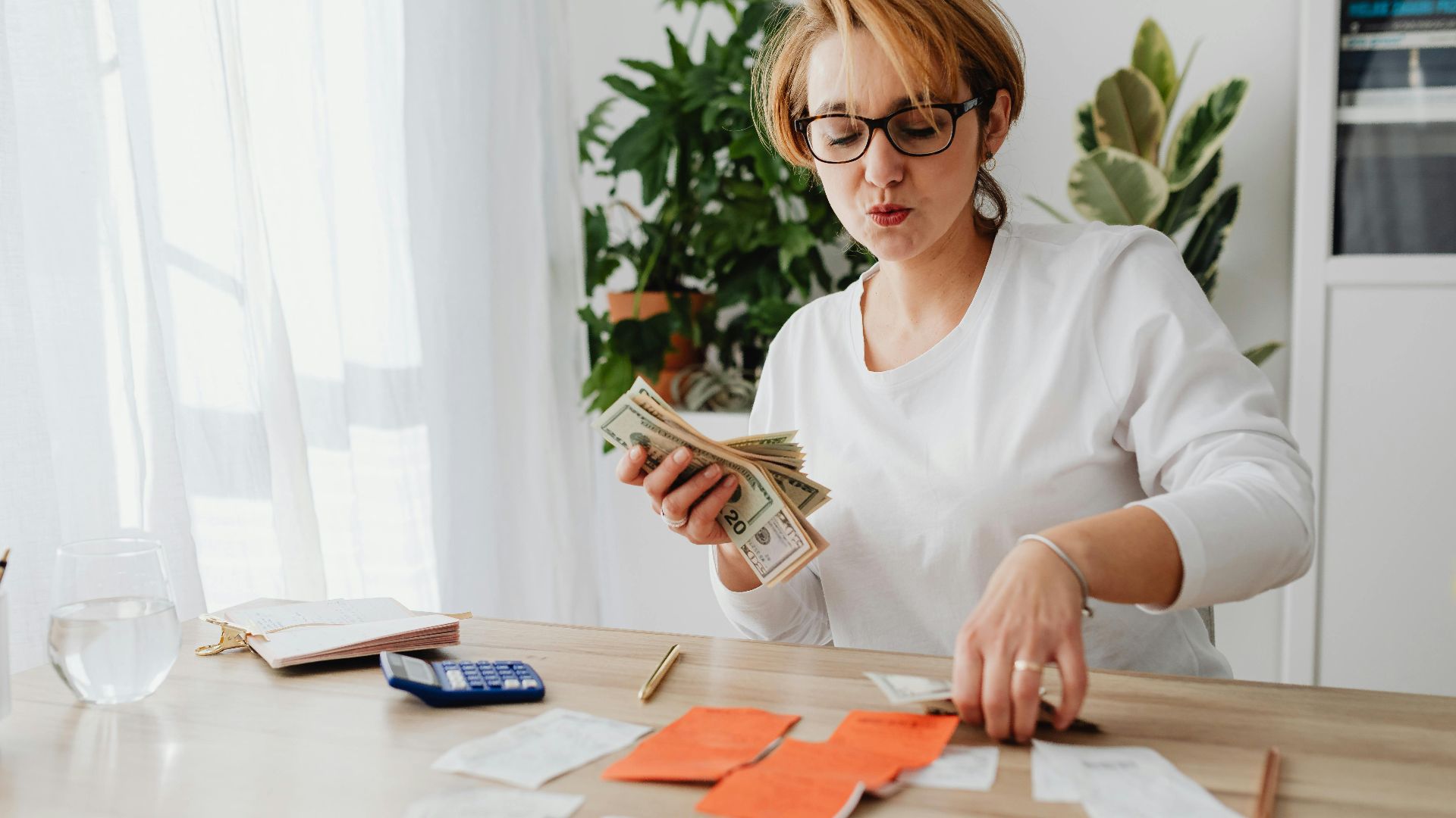 Woman sitting at a table counting cash and organizing receipts with a calculator nearby.