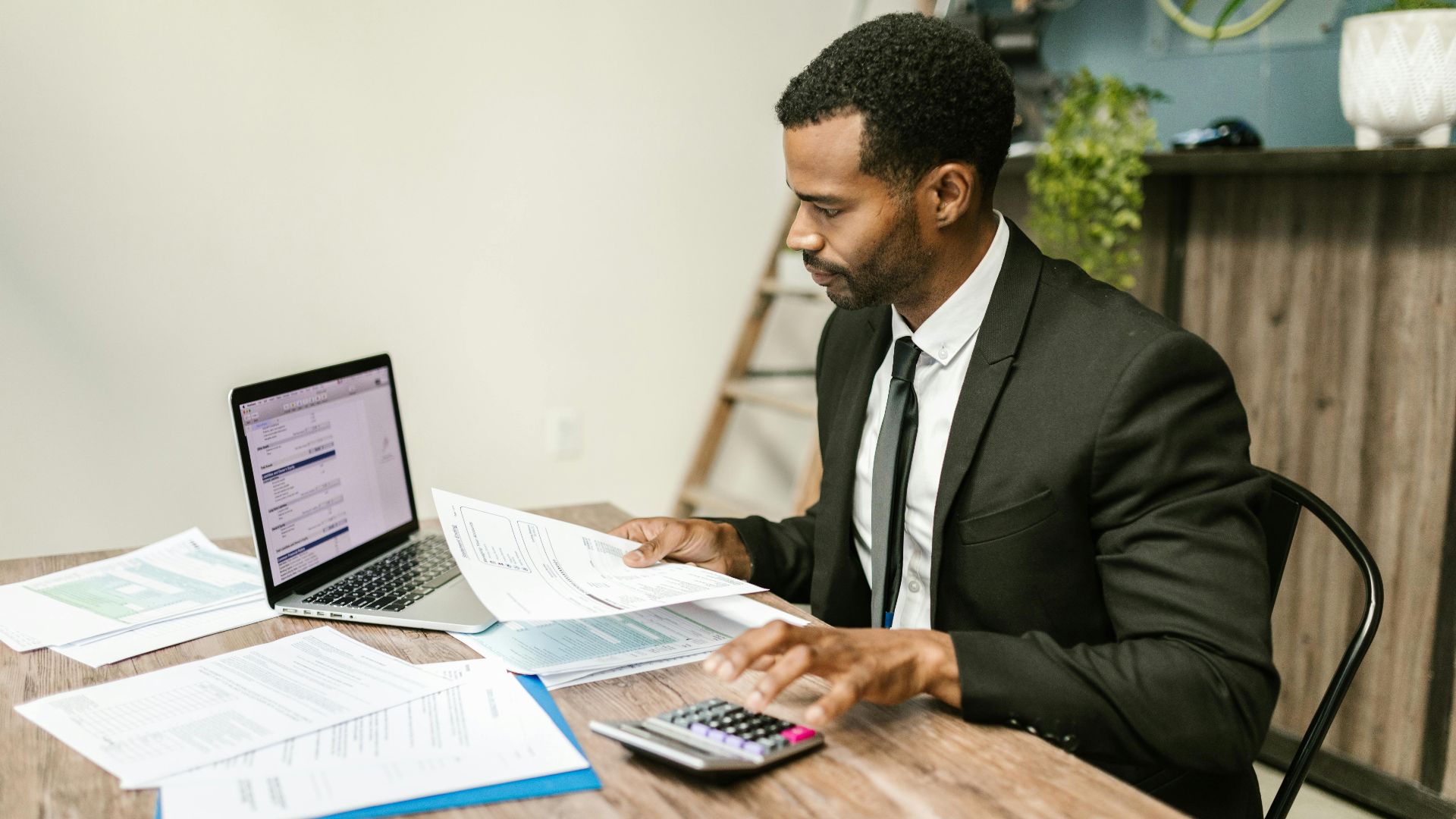 A businessman in a suit working on documents and using a laptop at an office desk.