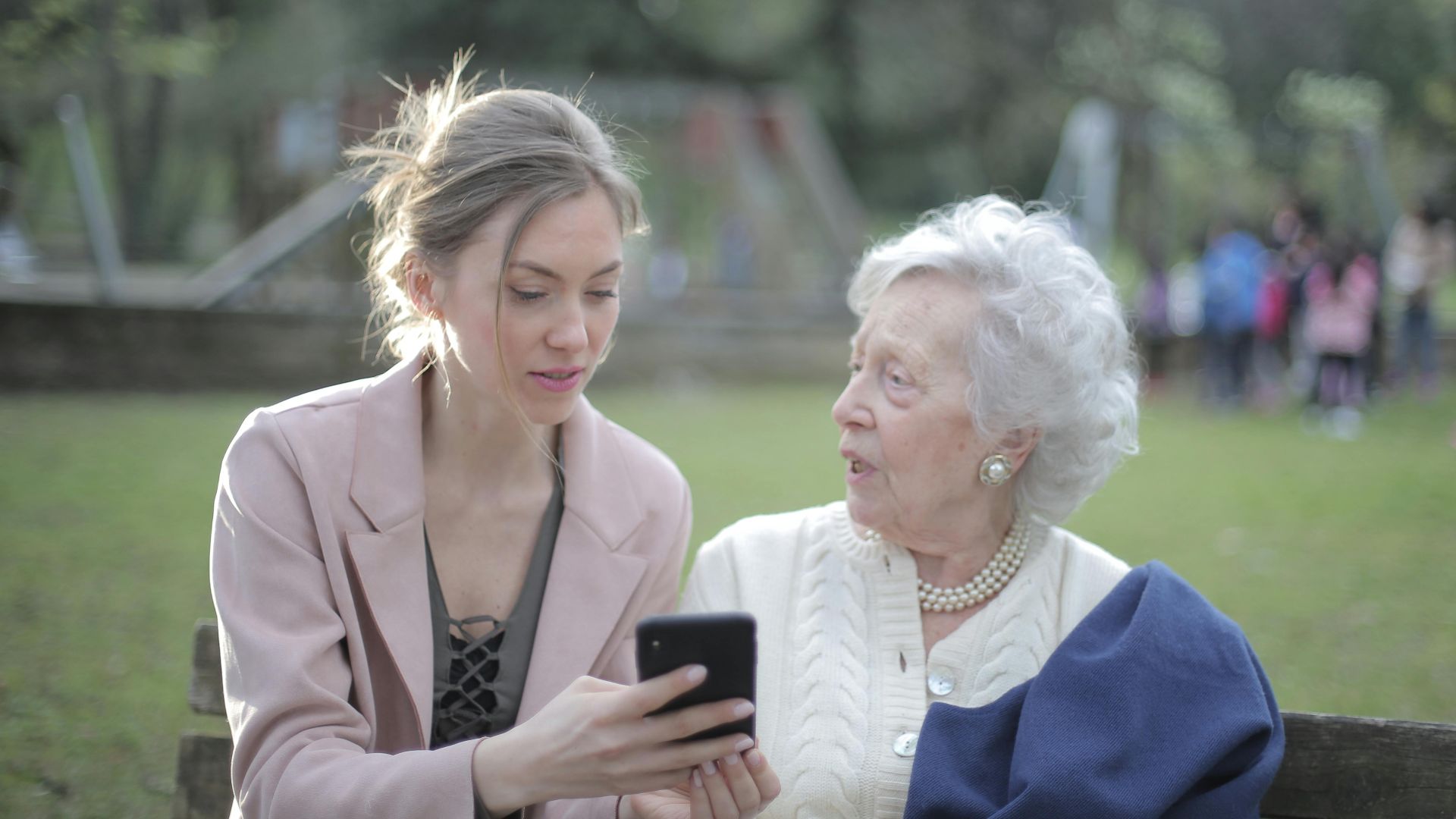 Concentrated female in elegant wear sitting on bench in park and instructing elderly mom on use of cellphone