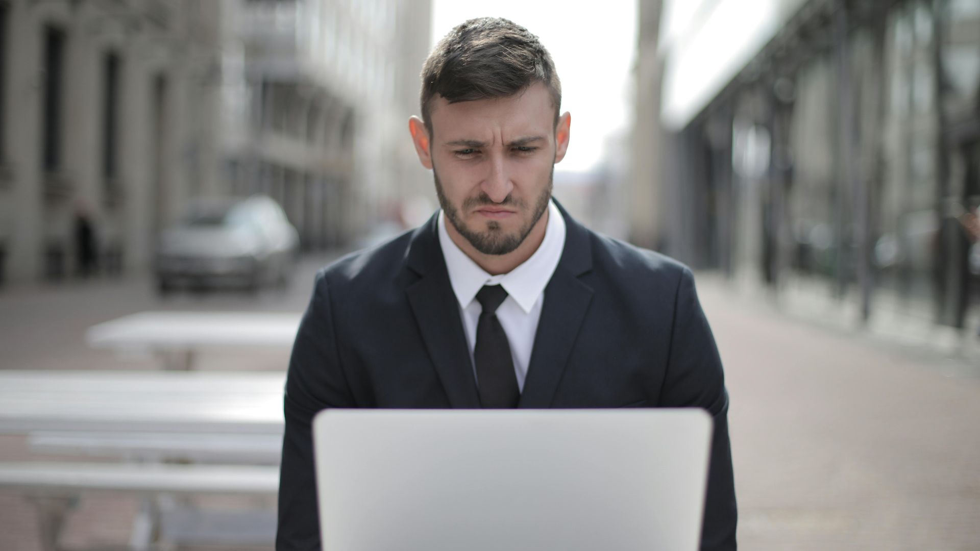 A focused businessman in a suit works on his laptop outdoors in an urban setting.