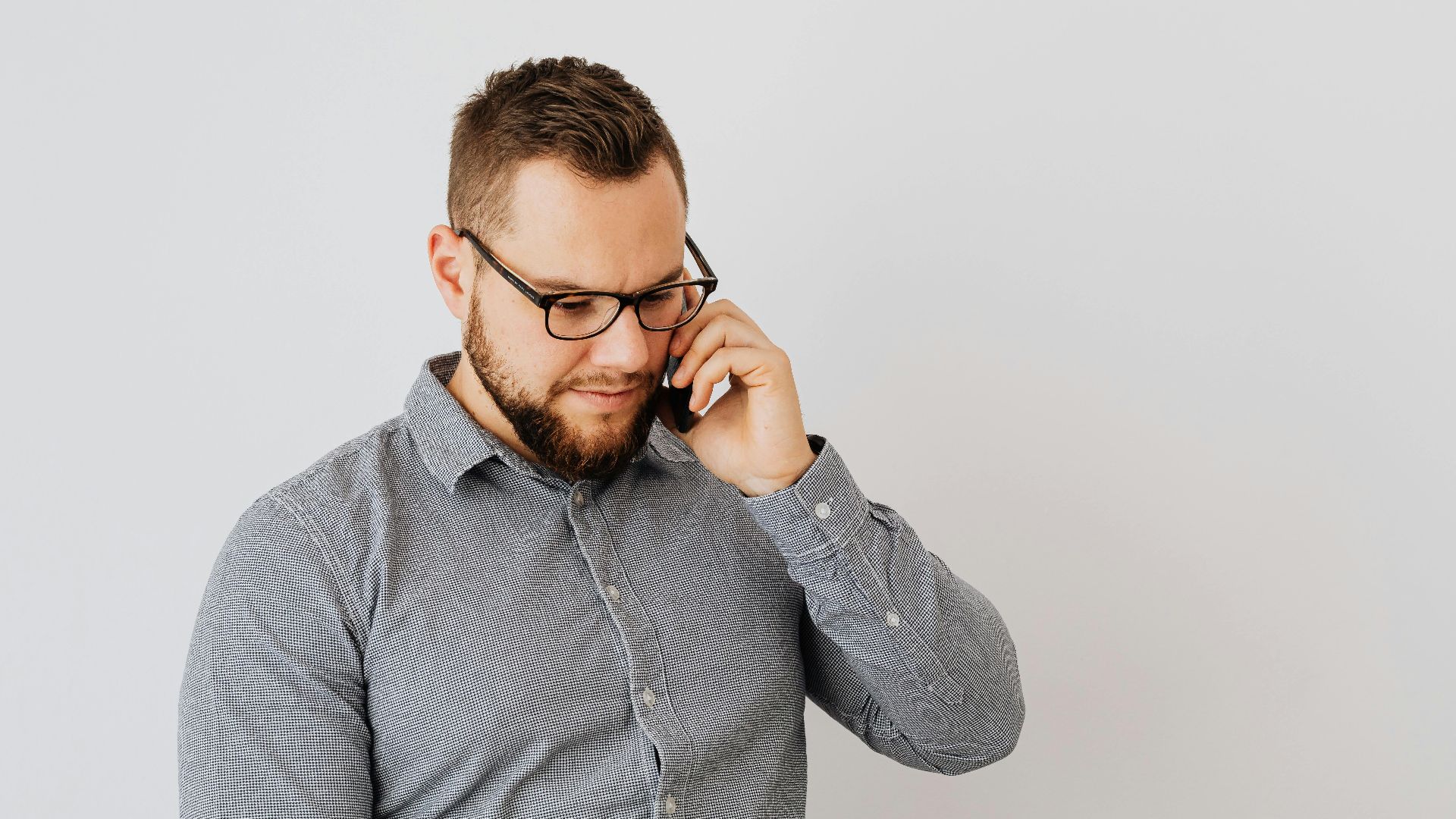Young man with beard in glasses and gray shirt talking on smartphone indoors.