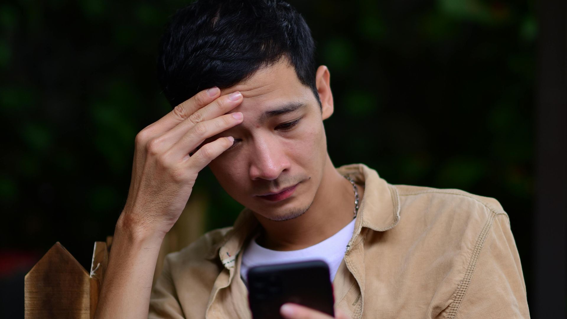 A young man looks stressed while checking his smartphone outdoors, possibly receiving bad news.