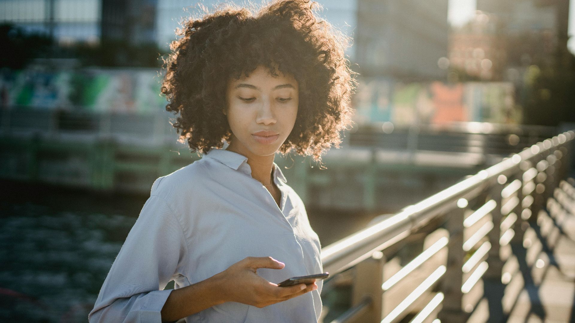 Portrait of a young woman with curly hair using her smartphone on an urban bridge during daylight.