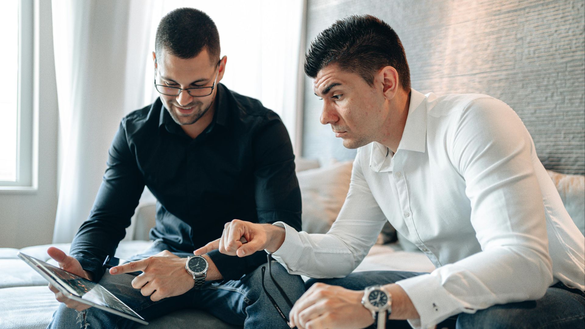 Two businessmen in casual clothing discuss a project on a tablet indoors.