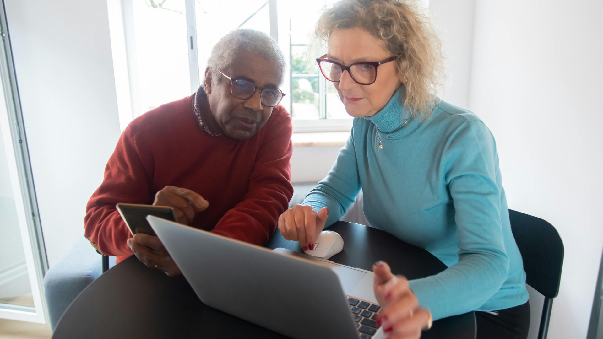 Elderly couple using a laptop in a bright room, focusing on online activities together.