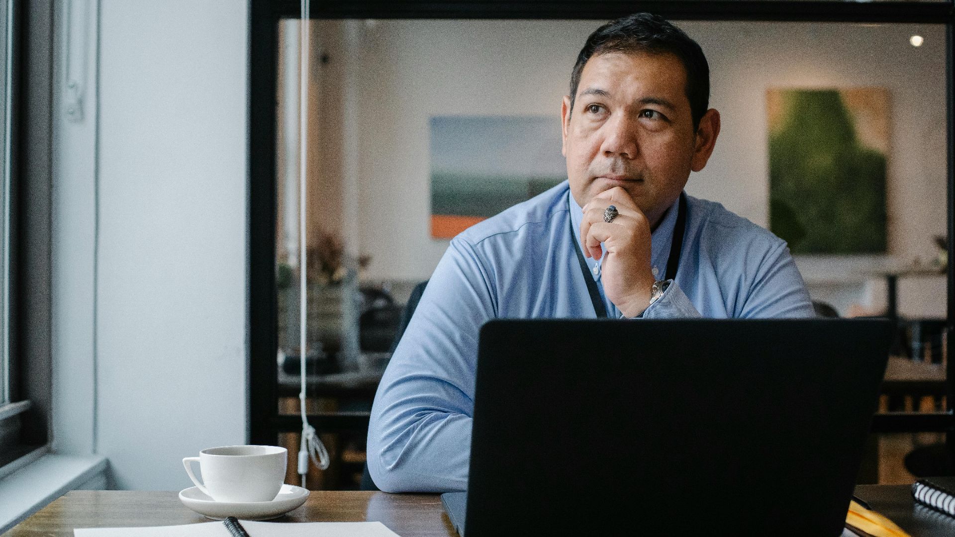 Concentrated ethnic adult male entrepreneur thinking on project while working at table with netbook and cup of drink