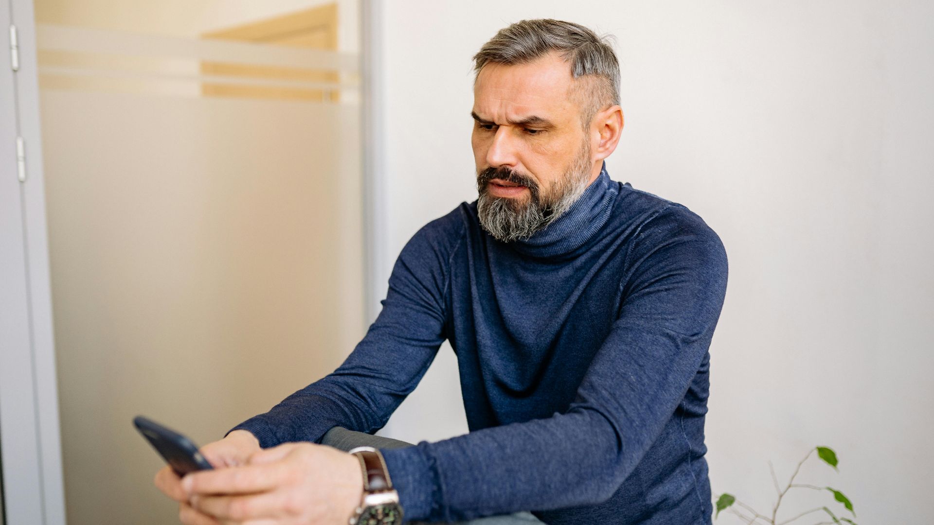 Serious middle-aged man sitting and using a smartphone indoors.