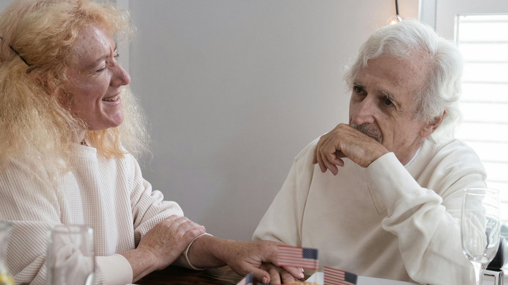 Elderly couple smiles and shares a moment over a meal with small American flags indoors.