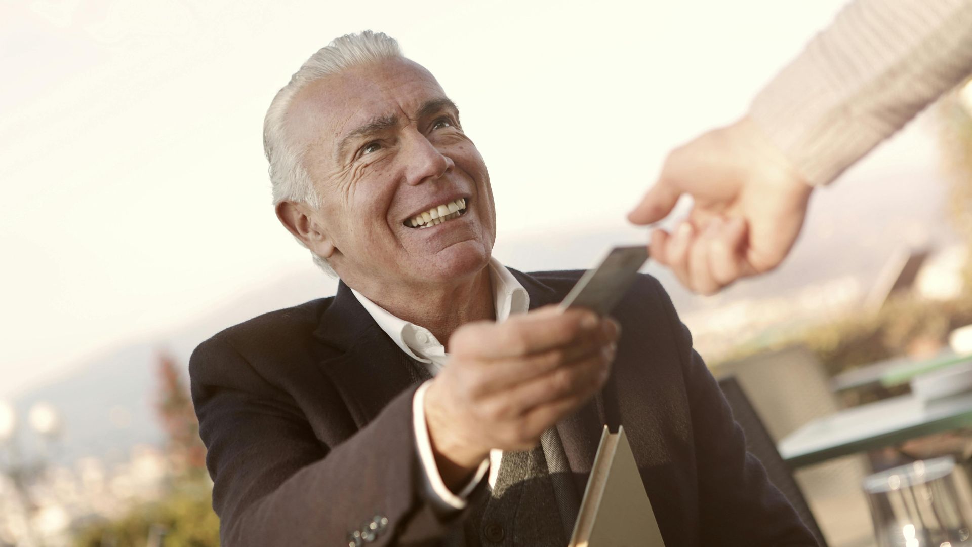 A senior man smiles as he hands a credit card outdoors, embodying trust and interaction.