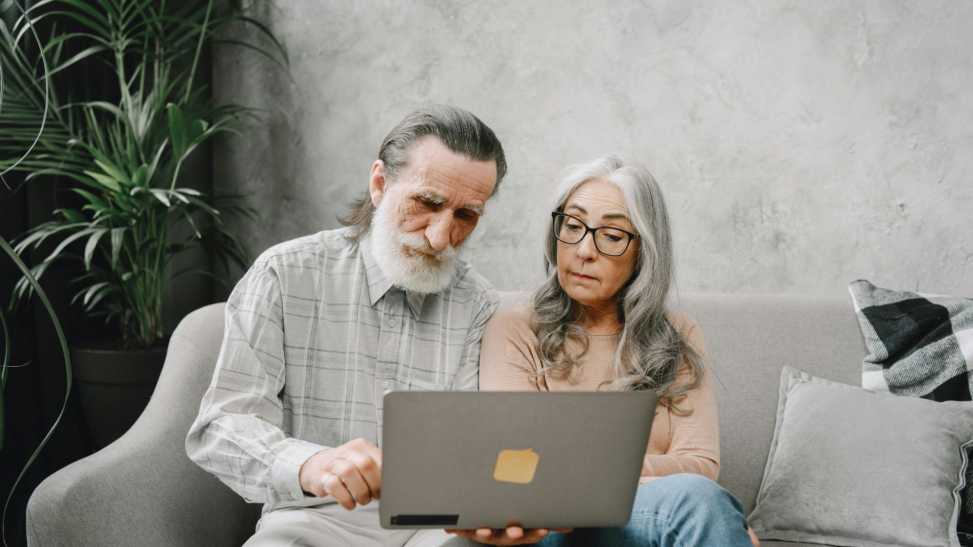 Senior couple sitting on a sofa using a laptop indoors, embracing technology together.