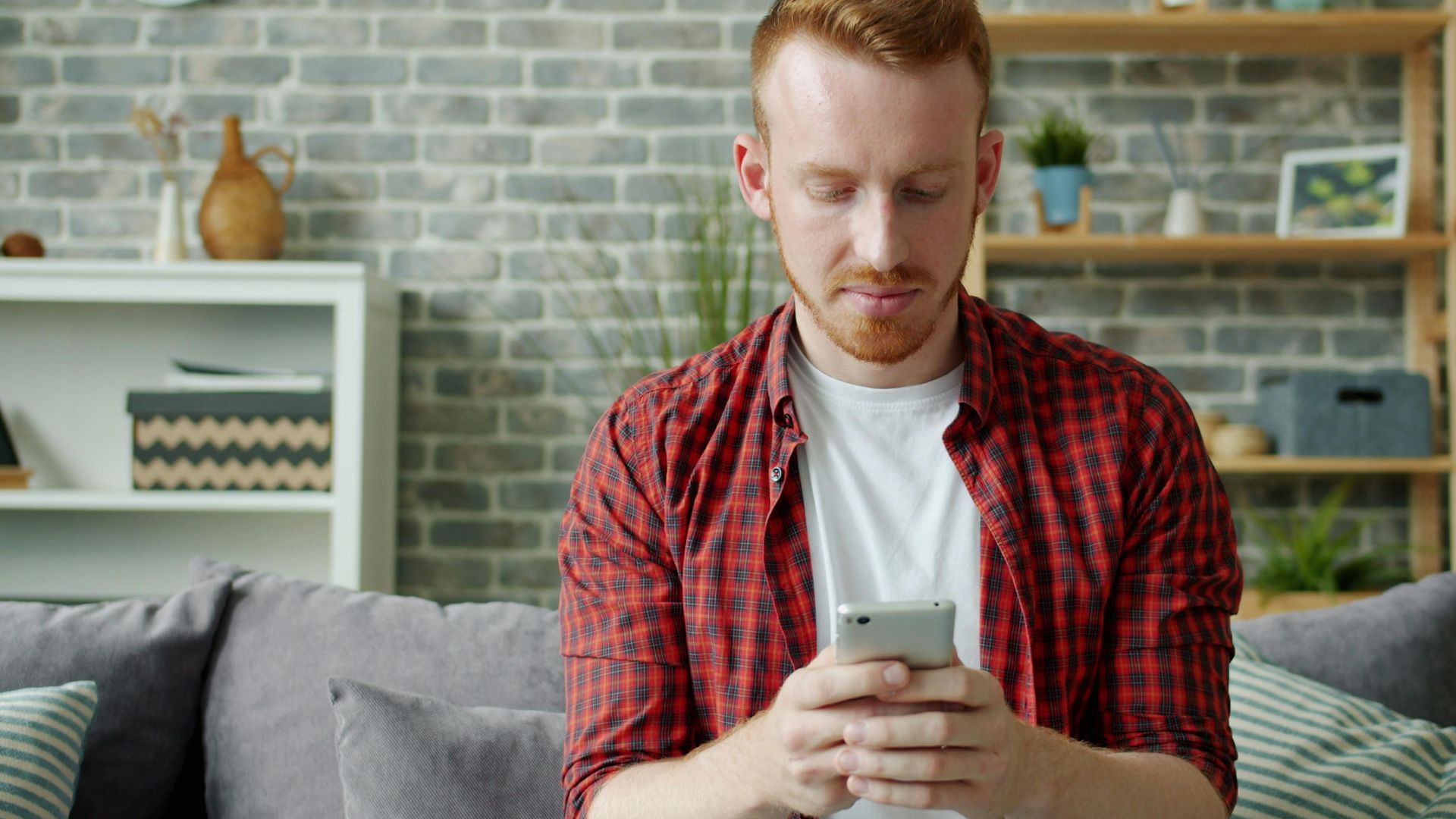 Man in red plaid shirt texting on smartphone at home, modern lifestyle.